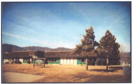 Exterior view of Evergreen School, a single-story school building with green-trimmed entryways and a flat roof, with large trees in front and rolling hills visible in the background.