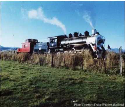 The Yreka Western Railroad steam locomotive with a red caboose car, traveling through a grassy field on the outskirts of Yreka with steam rising from the engine. Mountains are visible in the background under a partly cloudy sky. Photo courtesy Yreka Western Railroad.