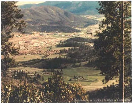 Panoramic view looking down from a hillside over the City of Yreka and the surrounding Shasta Valley. The city is nestled in the valley below, with forested ridgelines and mountains forming the backdrop. Pine trees frame the foreground. Photo courtesy Yreka Chamber of Commerce.